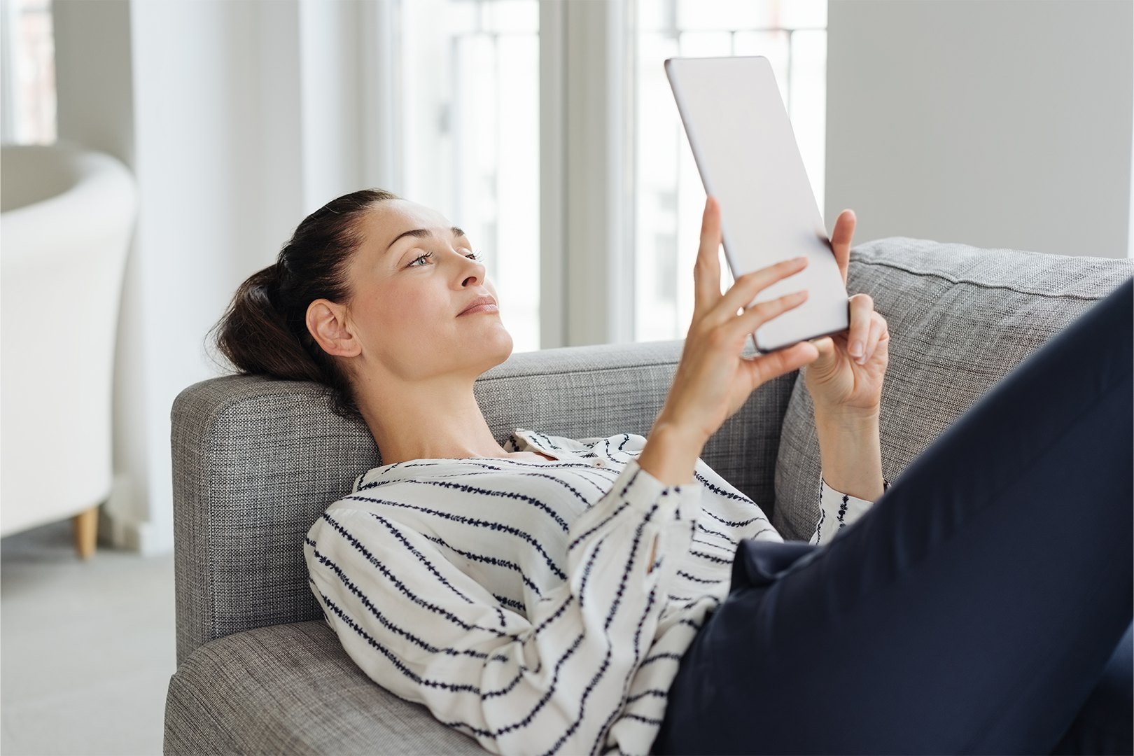 A woman in a striped shirt lying on a couch looking at a tablet